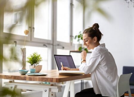 Eine Frau sitzt an einem Schreibtisch. Vor ihr ein Laptop und ein Notizbuch, in das sie etwas schreibt.