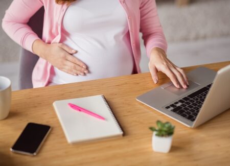 Eine schwangere Frau sitzt vor einem Schreibtisch. Vor sich einen Laptop und einen Notizblock.