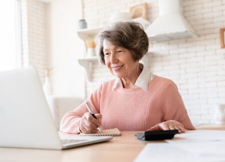 Eine ältere Frau sitzt vor einem Laptop. In der einen Hand hat sie einen Taschenrechner und in der anderen Hand einen Schreibblock.