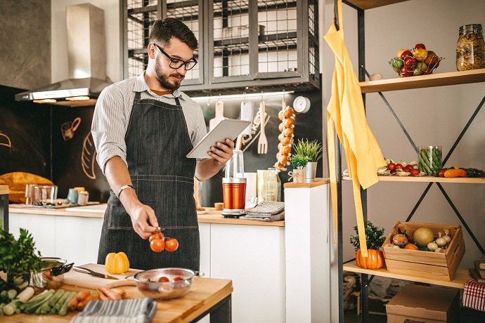 Ein Mann steht mit einem Tablet in der Hand in der Küche und bereitet ein Essen zu.