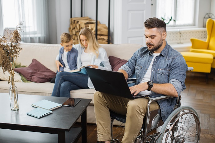 Ein Mann im Rollstuhl arbeitet am Laptop, während seine Frau und Sohn im Hintergrund auf dem Sofa sitzen